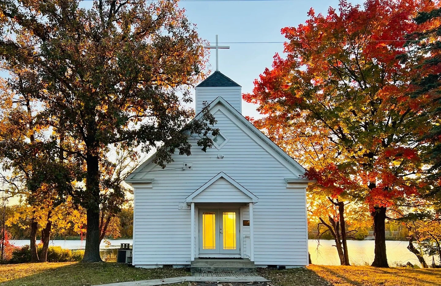 The white chapel at Legacy Lakeside Resort at golden hour — a cross above the steeple, its doors lit from inside, maples in full autumn color either side and the lake beyond