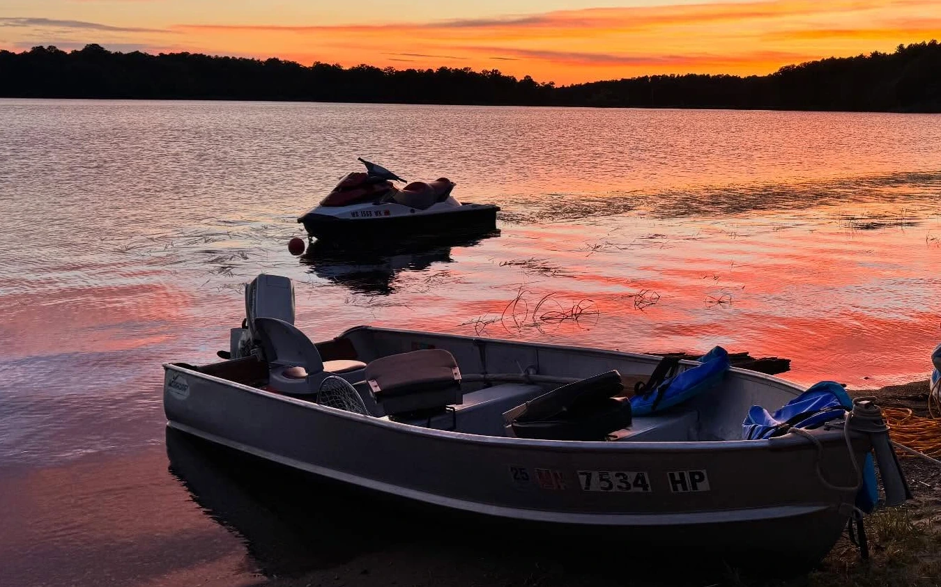 A fishing boat pulled up on the sand and a jet ski moored just offshore, with Spitzer Lake and the treeline under a red and orange sunset